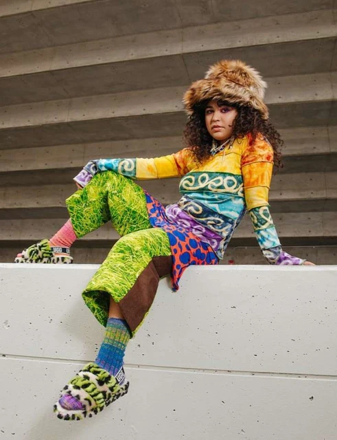 A stylish young woman in a colorful, patterned outfit and furry hat sits against a concrete wall, showcasing a bold fashion statement.