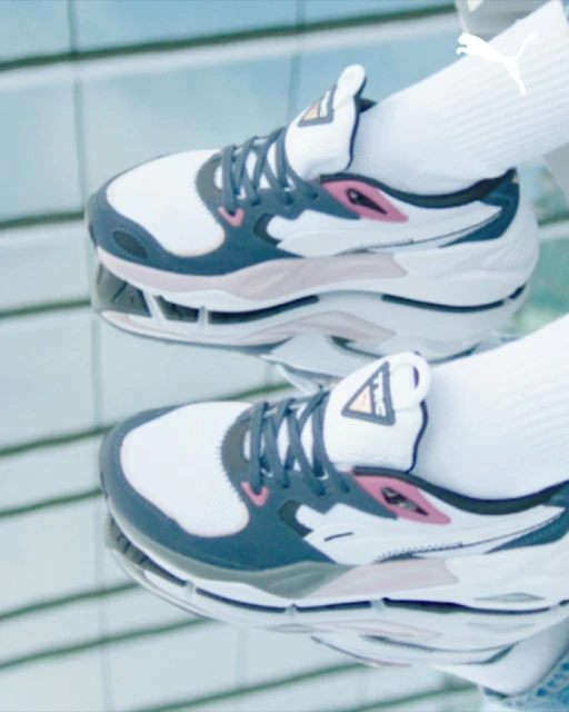 Close-up of a white, gray, and pink Puma sneaker with white sock, reflected in a mirror surface and featuring the Puma logo.