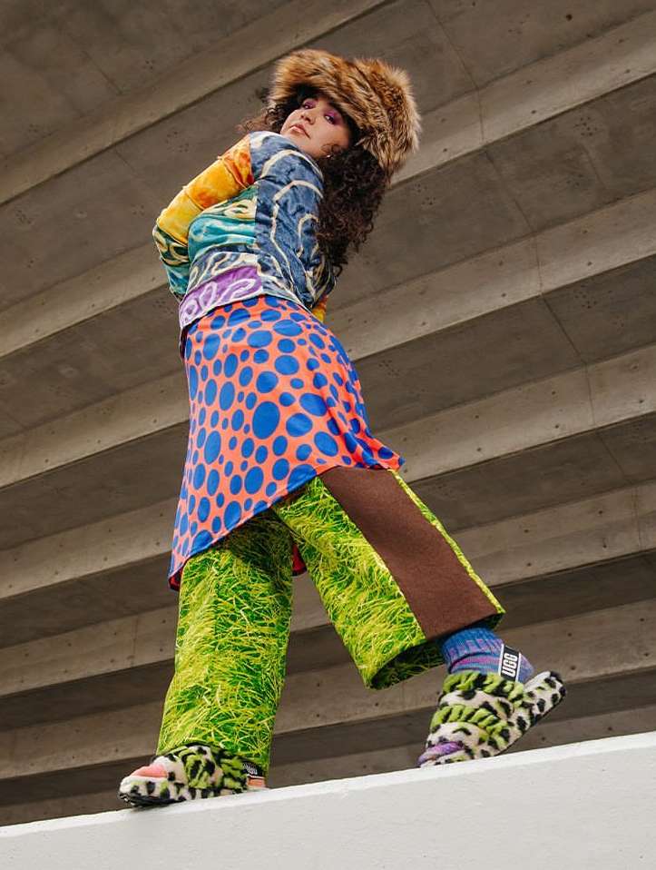 A young adult female fashion model poses in an eccentric outfit with bright colors and patterns in an urban setting, shot from a low angle.