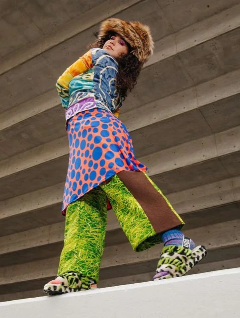 A young adult female fashion model poses in an eccentric outfit with bright colors and patterns in an urban setting, shot from a low angle.