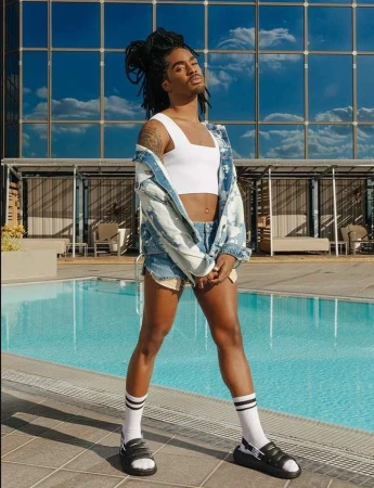 A person with dreadlocks wearing a white tank top, denim shorts, and slides by an outdoor pool.
