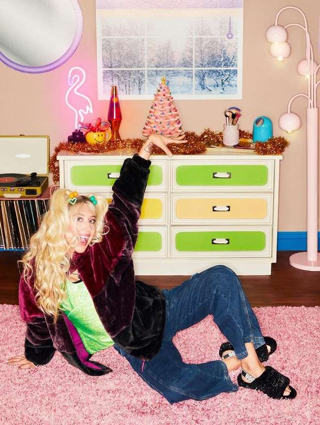 Blonde woman in retro outfit poses on pink carpet in a cozy room with Christmas tree, neon flamingo, and vintage decor accents.