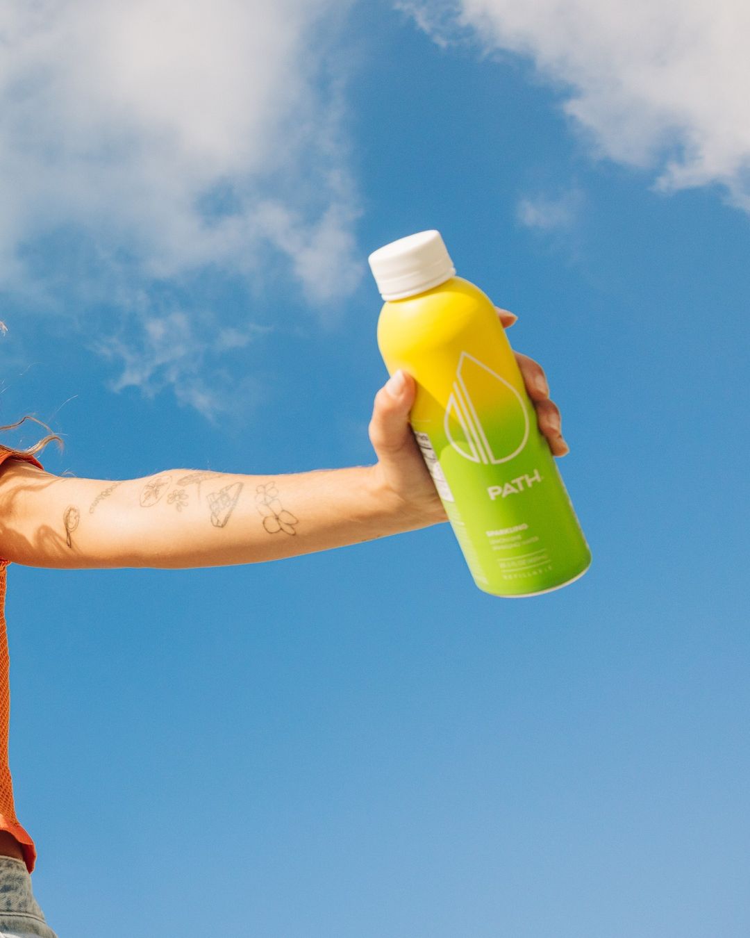 Adult hand holds a yellow aluminum bottle of PATH water against a vibrant blue sky with scattered clouds on a sunny day outdoors.