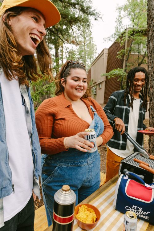 Three adults stand near a grill with beer and snacks in an outdoor setting, possibly a backyard or park, with trees in the background.