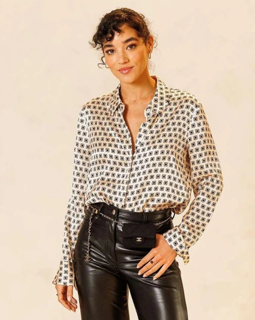 Portrait of a woman in a patterned blouse and leather pants with a Chanel belt bag, posing indoors against a neutral background in soft lighting.