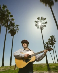 A man wearing a flat cap playing an acoustic guitar outdoors beneath towering palm trees against a clear blue sky
