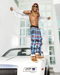 Lil Wayne posing energetically on a white car hood wearing plaid trousers and tan boots, photographed by Neil Favila