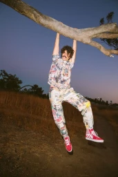 Man with curly hair and mustache hanging from a tree branch wearing patterned shirt, matching trousers, and red high-top sneakers at dusk.