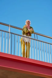 Woman wearing a pink hijab, olive green cardigan, and yellow trousers leaning against a metal railing under a clear blue sky.