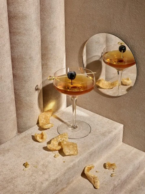 A cocktail on a stemmed glass with blueberry garnish, pork rinds, and a mirror, create a stylish drink and snack still life.
