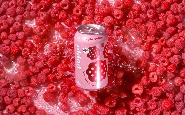 Pink Poppi Raspberry Rose can surrounded by fresh raspberries in a watery setting, showcasing product and flavour ingredients.