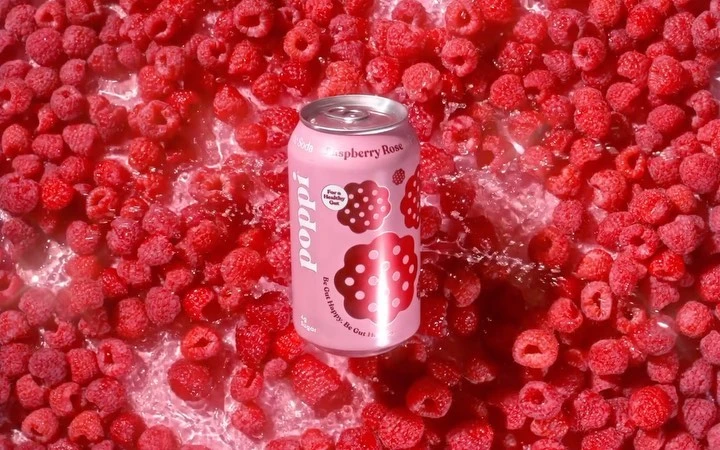 Pink Poppi Raspberry Rose can surrounded by fresh raspberries in a watery setting, showcasing product and flavour ingredients.