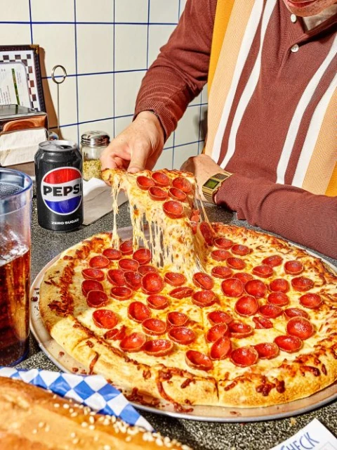 Overhead medium shot of a person taking a slice of pepperoni pizza with melting cheese, with a can of Pepsi, and a baguette.