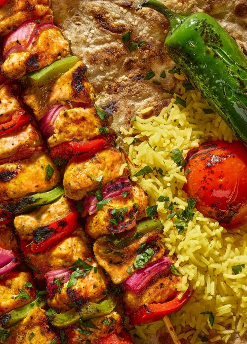 Overhead shot of chicken skewers with red onion and bell peppers served with pita bread, rice, and a grilled tomato and pepper.