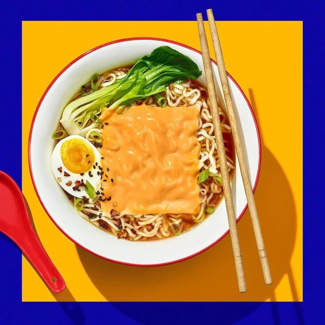 Overhead shot of ramen in a white bowl with chopsticks, boiled egg, bok choy, and orange topping on a yellow and blue background.