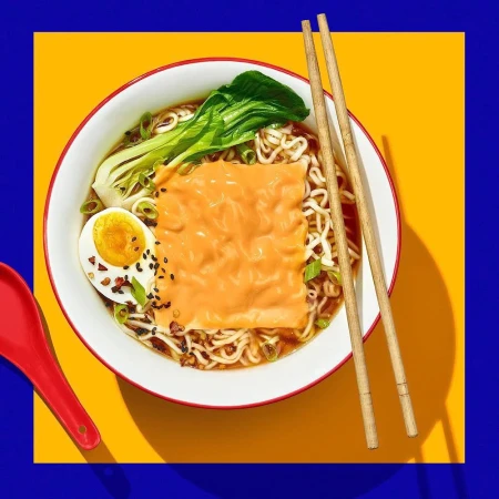 Overhead shot of ramen in a white bowl with chopsticks, boiled egg, bok choy, and orange topping on a yellow and blue background.
