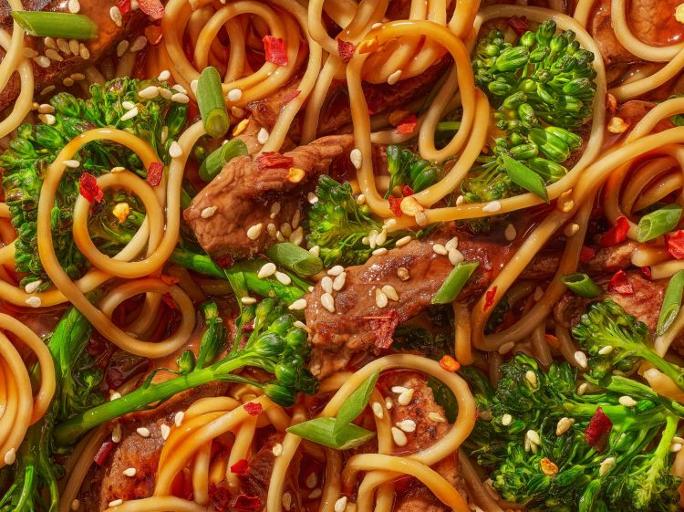 Overhead close-up of stir-fried noodles with broccoli and meat, topped with sesame seeds and red pepper flakes, served as a savory dish.