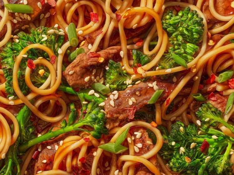Overhead close-up of stir-fried noodles with broccoli and meat, topped with sesame seeds and red pepper flakes, served as a savory dish.