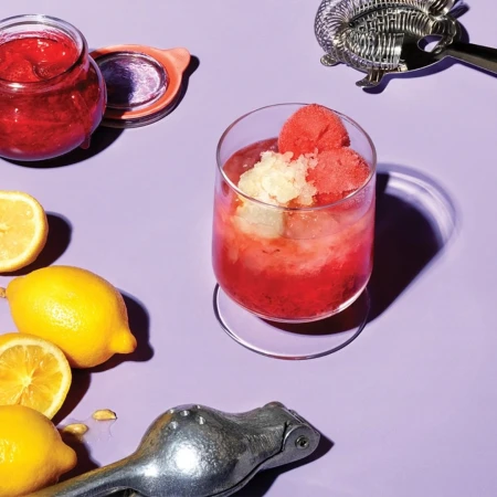 Cocktail in a glass with lemons, a jar of jam, and bar tools on a lavender background, highlighting food and beverage photography.