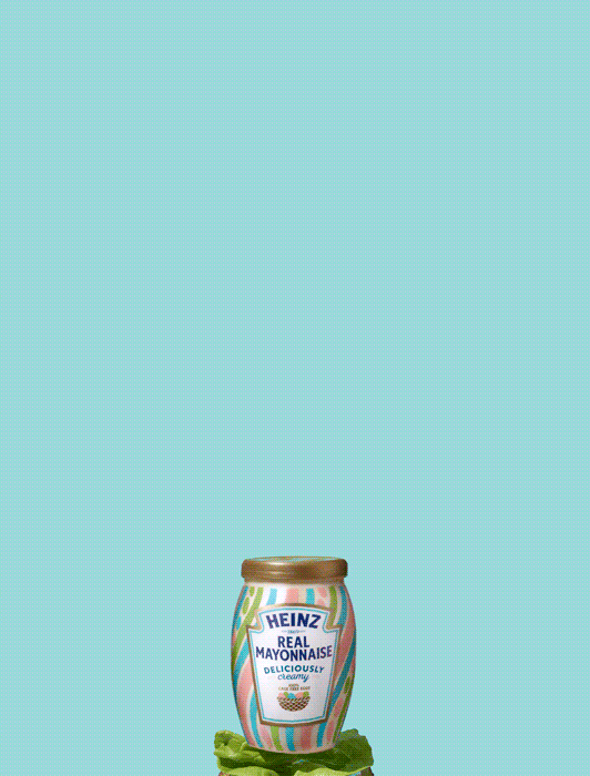 A jar of Heinz Real Mayonnaise with colorful stripes sits atop green lettuce against a solid light blue background.