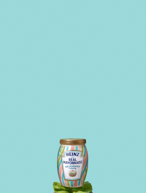 A jar of Heinz Real Mayonnaise with colorful stripes sits atop green lettuce against a solid light blue background.