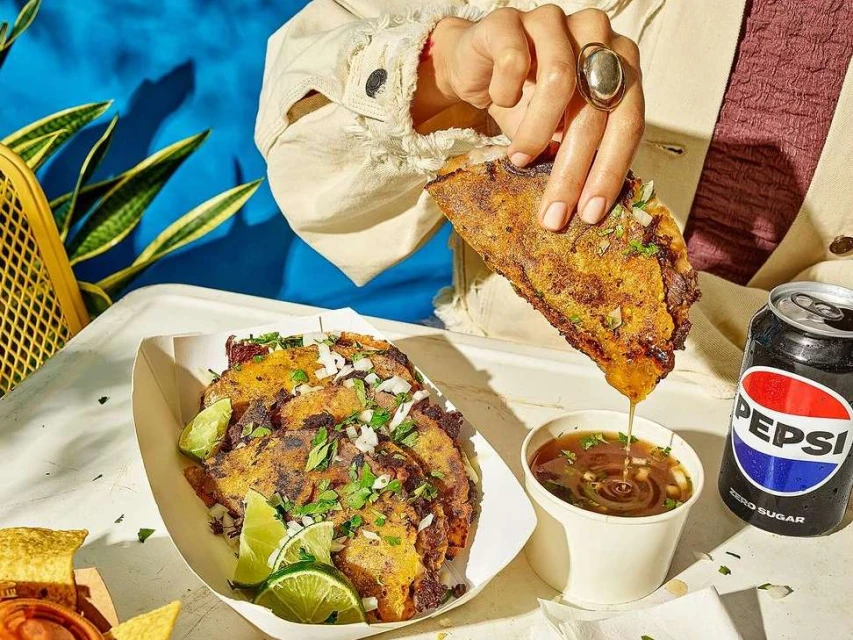 Close-up of crispy birria tacos, garnished with onions and cilantro, being dipped in broth, with a can of Pepsi and blue background.