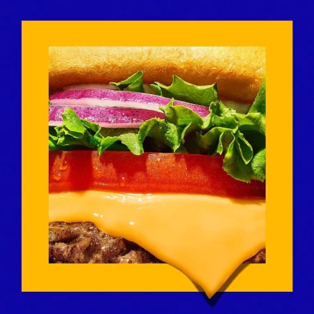 Extreme close-up of a stacked burger showing lettuce, tomato, red onion and cheese on a blue background with a yellow square border.