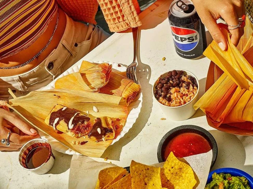 Overhead shot of a picnic table laden with various foods, including tamales, chips, guacamole, salsa, rice, beans, and a can of Pepsi.