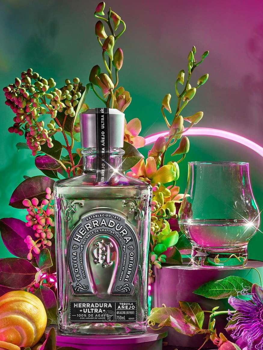 A bottle of Herradura tequila is showcased alongside a glass, set against a vibrant backdrop of flowers, foliage, and neon lighting.