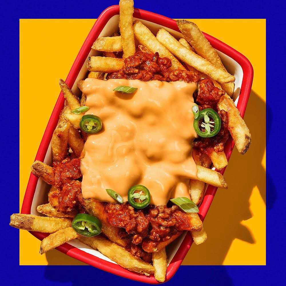 Overhead shot of chili cheese fries in red dish with yellow and blue background. French fries covered in chili, cheese sauce, jalapenos.