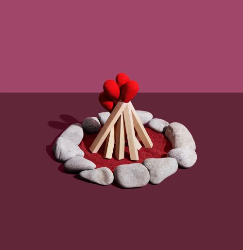 Stylized campfire made of oversized matchsticks, arranged in a teepee formation on a red surface, surrounded by grey rocks.