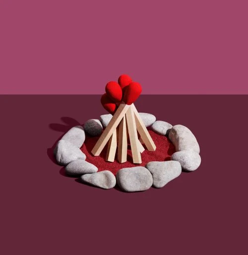 Stylized campfire made of oversized matchsticks, arranged in a teepee formation on a red surface, surrounded by grey rocks.