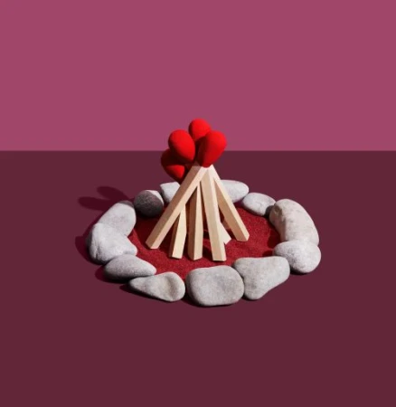 Stylized campfire made of oversized matchsticks, arranged in a teepee formation on a red surface, surrounded by grey rocks.
