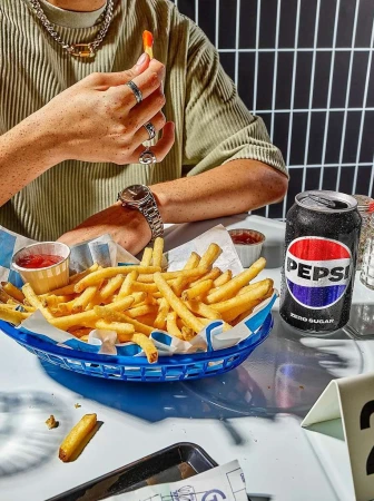 Person eating french fries from a blue basket, next to a Pepsi Zero Sugar can, in a brightly lit indoor setting.