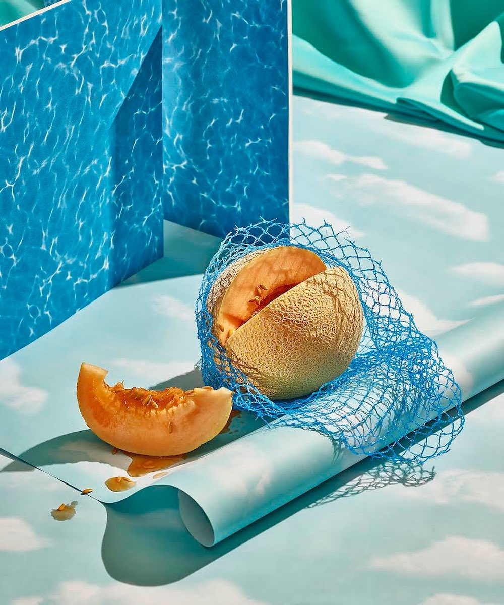 Still life food photography of cantaloupe melon, slice, and blue netting set against light blue background and water reflection backdrop.