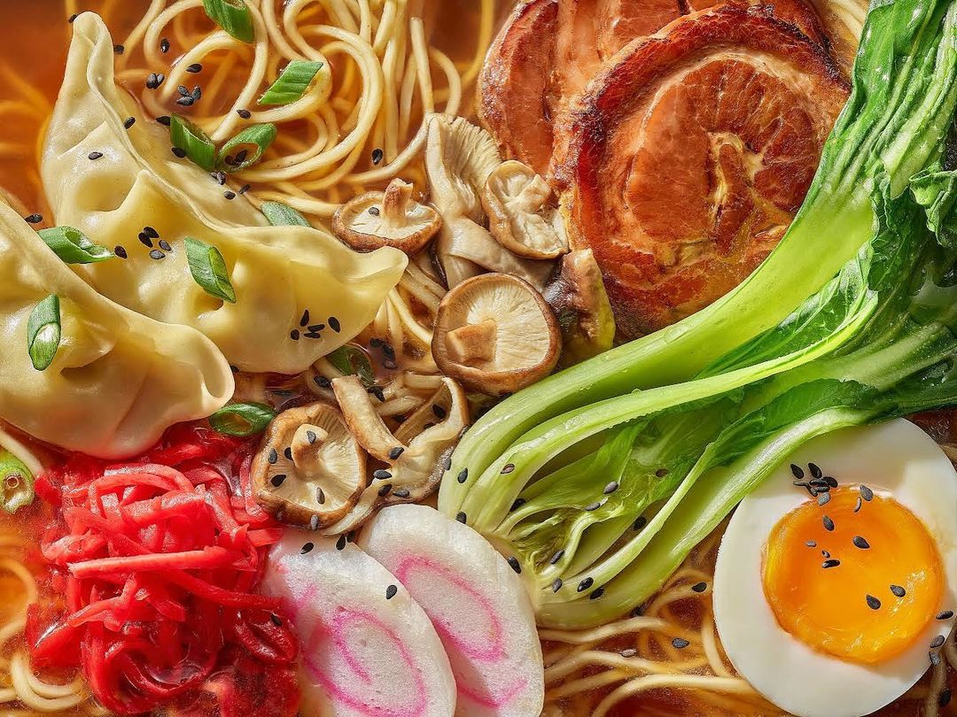 Overhead shot of a flavourful ramen bowl featuring noodles, pork, bok choy, dumplings, egg, mushrooms, scallions and bright red ginger.