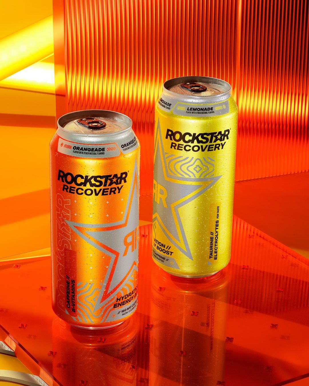 Two cans of Rockstar Recovery energy drinks, one orangeade and one lemonade flavor, stand on a reflective orange surface in a product shot.