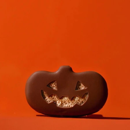 Halloween treat: chocolate-covered pumpkin-shaped candy with jack-o'-lantern face on an orange background, festive and delicious.