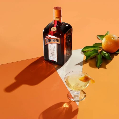Cointreau bottle, cocktail glass, and orange on a vibrant orange and white surface, showcasing beverage presentation.