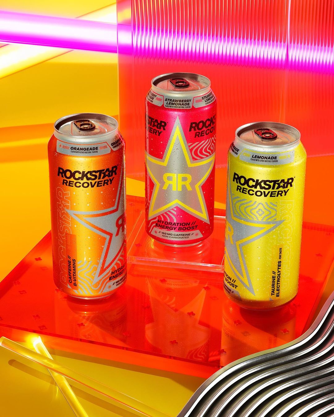 Three cans of Rockstar Recovery energy drink in orange, red, and yellow, arranged on an orange reflective surface in a bright studio setting.