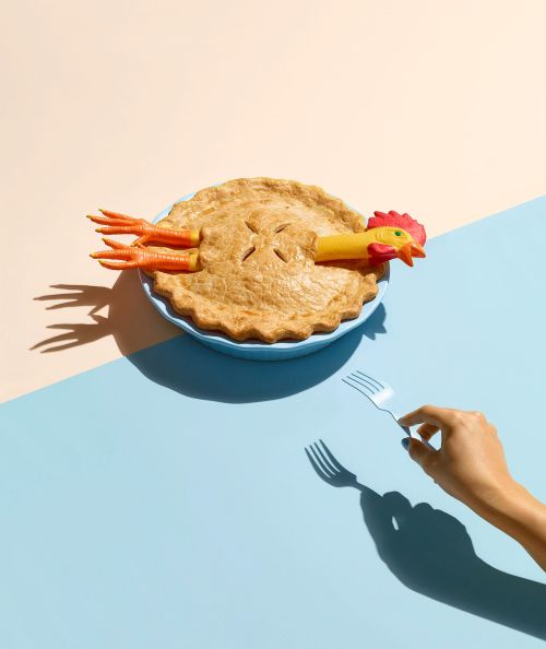 Novel conceptual image of a pie in a blue dish with a rubber chicken inside and a hand holding a fork against a two-tone background.