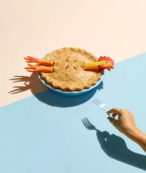 Novel conceptual image of a pie in a blue dish with a rubber chicken inside and a hand holding a fork against a two-tone background.