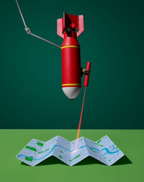 A red bomb with a robotic arm directs a red laser at a folded map against a green backdrop, creating a visually striking and intriguing still life.