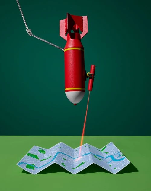 A red bomb with a robotic arm directs a red laser at a folded map against a green backdrop, creating a visually striking and intriguing still life.