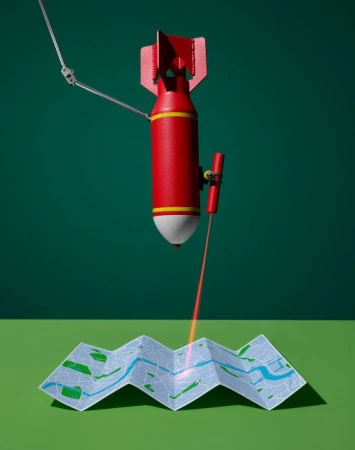 A red bomb with a robotic arm directs a red laser at a folded map against a green backdrop, creating a visually striking and intriguing still life.