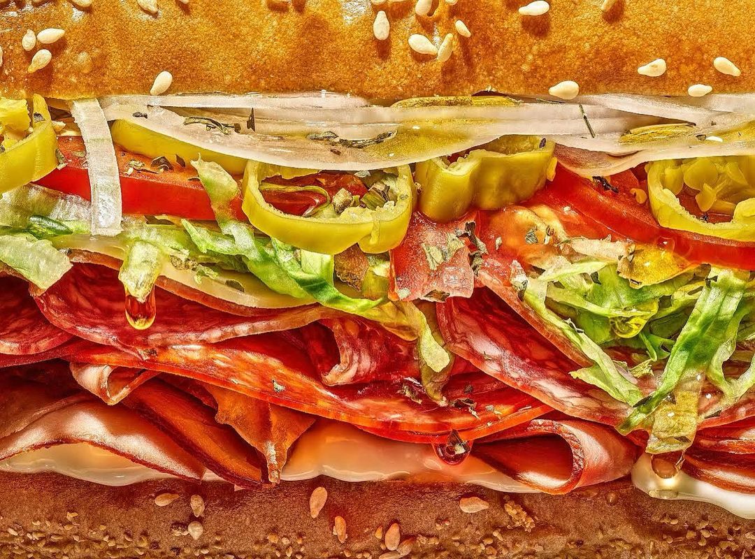 Close-up of a stacked sandwich showcasing layers of salami, lettuce, tomatoes, onions, and peppers, and a sesame seed bun.