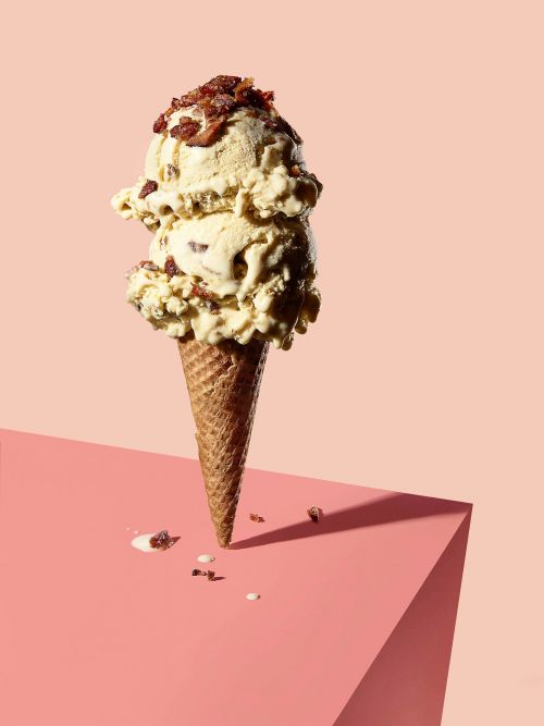 Ice cream cone with two scoops of bacon ice cream topped with bacon bits on a pink geometric surface and light pink background.