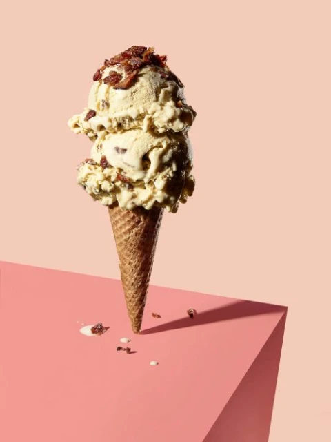 Ice cream cone with two scoops of bacon ice cream topped with bacon bits on a pink geometric surface and light pink background.