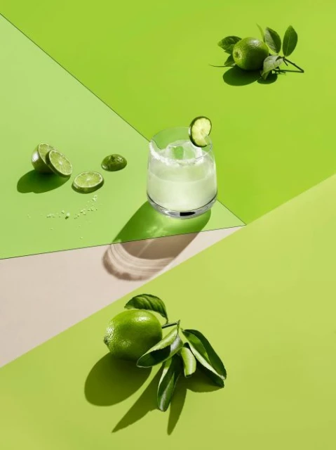 Still life shot of a cocktail in a glass with fresh limes, set against a geometric background in various shades of green and beige.