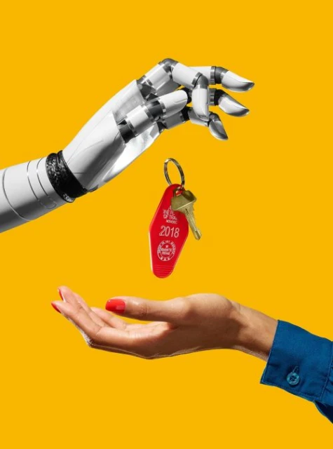 Robot hand offers a key on a red motel keychain to a human hand against a bright yellow background, showcasing technology and hospitality.